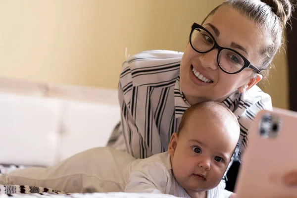 Childhood, motherhood, family concepts - successful smiling rich business woman Lady mother sit on bed with infant child baby talking on phone by video call, take photo at home in distance indoors