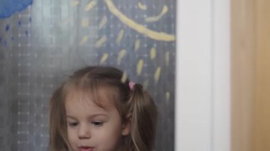 Çocukluk, yaratıcılık, doğa, ilkbahar, yaz konsepti - 3-4 yaşındaki sevimli küçük kızın yakın çekimi pencere camında akrilik renkler çiziyor. Anaokulu resim manzarası: güneş, ağaç, bulut, yağmur, çiçek