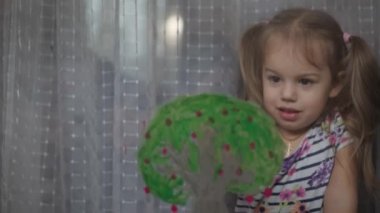 Çocukluk, yaratıcılık, doğa, ilkbahar, yaz konsepti - 3-4 yaşındaki sevimli küçük kızın yakın çekimi pencere camında akrilik renkler çiziyor. Anaokulu resim manzarası: güneş, ağaç, bulut, yağmur, çiçek