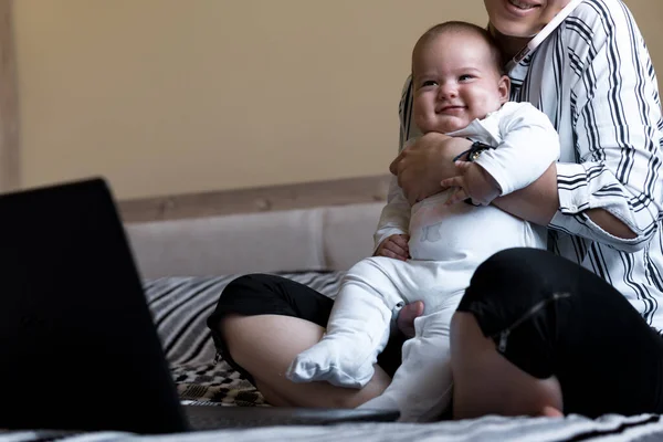 Childhood, motherhood, family concepts - successful smiling rich business woman Lady mother sit on bed with infant child baby talking on phone working on lap top computer at home in distance indoors