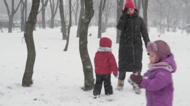 Kış, tatil, oyunlar, aile kavramları - iki mutlu anaokulu çocuğu, anneleriyle birlikte şapka ve eldiven takmış çocuklar, soğuk mevsimde kartopu oynuyorlar.