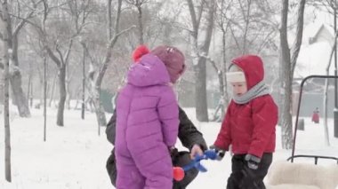 Kış, tatil, oyunlar, aile kavramları - iki mutlu anaokulu çocuğu, anneleriyle birlikte şapka ve eldiven takmış çocuklar, parkta soğuk mevsimde kartopu oynuyorlar.