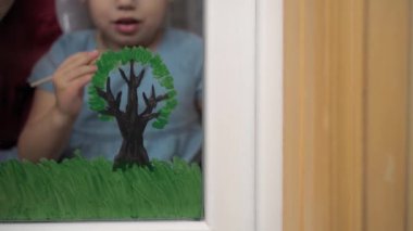 Çocukluk, annelik, yaratıcılık, doğa, sanat, bahar, yaz konsepti - 3-4 yaşlarında küçük kızı olan genç bir annenin pencere camına çizimi. kadın ve anaokulu kızı ağaç boyuyorlar..