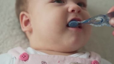 Bebek, çocukluk, hijyen konsepti, gülümseyen mutlu, tombul suratlı küçük çocuğun kendine gelmesi. Annem ilk kez bebek dişlerini mavi diş fırçasıyla fırçalıyor. Beyaz yumuşak yatakta 7 ay bebeğim.