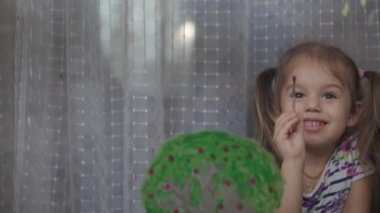 Çocukluk, yaratıcılık, doğa, ilkbahar, yaz konsepti - 3-4 yaşındaki sevimli küçük kızın yakın çekimi pencere camında akrilik renkler çiziyor. Anaokulu resim manzarası: güneş, ağaç, bulut, yağmur, çiçek