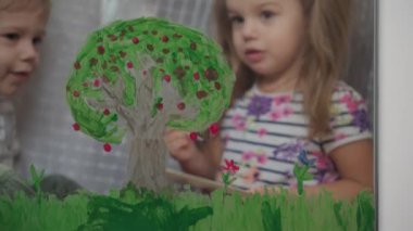 Çocukluk, yaratıcılık, doğa, ilkbahar, yaz konsepti - pencere camına akrilik renk çizen iki küçük anaokulu çocuğunun yakın çekimi. Kardeşlerin çocukları ve kız kardeşlerin manzara resimleri: ağaç, çimen