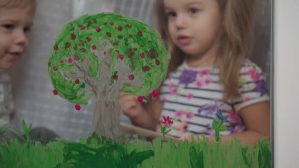 Dzieciństwo, kreatywność, natura, wiosenna, letnia koncepcja - zbliżenie dwójki małych przedszkolaków rysujących akrylowym kolorem na szybie okna. rodzeństwo brat z siostra malować krajobraz: drzewo, trawa — Wideo stockowe