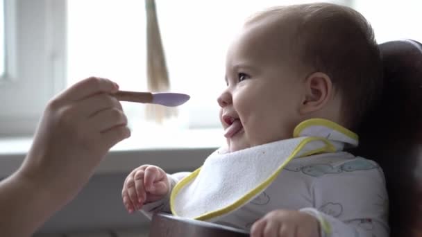 Питание, питание, детские концепции - мама кормит младенца резиновой ложкой. Симпатичный неголодный ребенок, одетый в биб, сидит в детском кресле, не хочет есть блюдо на кухне в помещении. не нравится первое кормление — стоковое видео