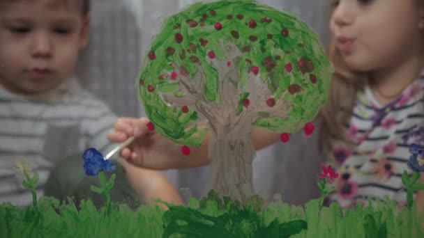 La infancia, la creatividad, la naturaleza, la primavera, el concepto veraniego - el primer plano de dos pequeños niños preescolares dibujan con el color acrílico en el cristal de la ventana. hermano niños hermano con hermana pintura paisaje: árbol, hierba — Vídeo de stock