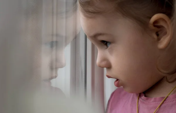 Kindertijd, eenzaamheid, isolatie, verdriet, hoop concepten - Een verdrietig schattig bezorgd Peuter baby meisje in roze met paardenstaarten kapsel kijkt door het raam. reflectie van het gezicht van het kleine lieve kind — Stockfoto