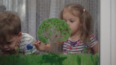Çocukluk, yaratıcılık, doğa, ilkbahar, yaz konsepti - pencere camına akrilik renk çizen iki küçük anaokulu çocuğunun yakın çekimi. Kardeşlerin çocukları ve kız kardeşlerin manzara resimleri: ağaç, çimen