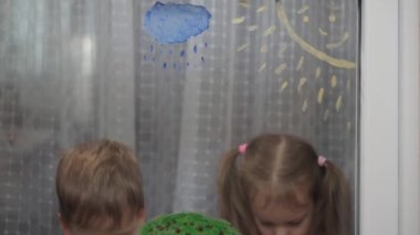 Çocukluk, yaratıcılık, doğa, ilkbahar, yaz konsepti - pencere camına akrilik renk çizen iki küçük anaokulu çocuğunun yakın çekimi. Kardeşlerin çocukları ve kız kardeşlerin manzara resimleri: ağaç, çimen