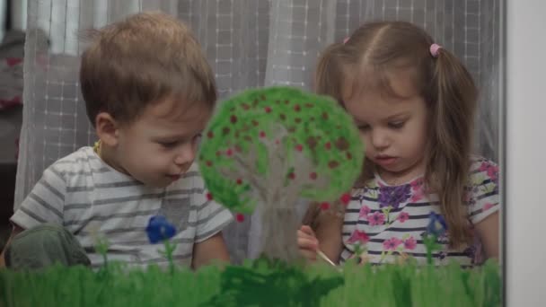 Дитинство, творчість, природа, весна, літня концепція крупним планом двох маленьких дітей дошкільного віку малюють акриловим кольором на склі вікна. дітей-братів з сестринською фарбою пейзаж: дерево, трава — стокове відео