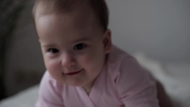 Bebé, concepto de la infancia - primer plano de sonriente feliz regordeta divertida cara gordito de ojos marrones del niño niña niña despierta. 9-12 meses bebé muecas, cierra los ojos se arrastra sentarse en la cama suave de color rosa en casa en interiores — Vídeo de stock