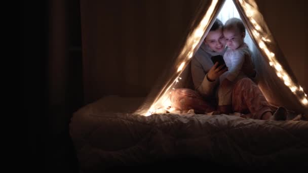 Authentic cute mom with chubby babe infant girl in tent at home. young woman use smartphone with kid 6-12 months old sit in wigwam at night. Family, Childhood, Motherhood, Comfort and Safety concept. — Stock Video