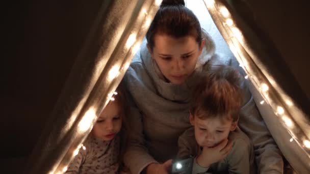 Auténtica tierna mamá linda y niños preescolares niño y niña en la tienda de campaña en casa. joven mujer leer libro a los niños 2-4 años de edad en wigwam por la noche. Familia, Infancia, Maternidad, Confort y Seguridad — Vídeos de Stock