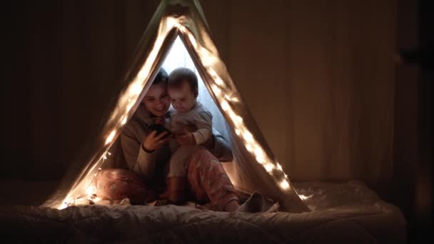 Authentieke schattige moeder met mollig babe baby meisje in tent thuis. jonge vrouw gebruik smartphone met kind 6-12 maanden oud zitten in wigwam 's nachts. Familie, Jeugd, Moederschap, Comfort en Veiligheid. — Stockvideo