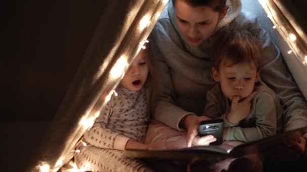 Auténtica tierna mamá linda y niños preescolares niño y niña en la tienda de campaña en casa. joven mujer leer libro a los niños 2-4 años de edad en wigwam por la noche. Familia, Infancia, Maternidad, Confort y Seguridad — Vídeo de stock