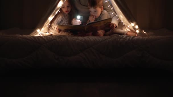 Autentické něžné roztomilé maminka a školka děti chlapec a dívka ve stanu doma. mladá žena číst knihu pro děti 2-4 roky ve vigvamu v noci. Rodina, dětství, mateřství, pohodlí a bezpečnost koncepce — Stock video