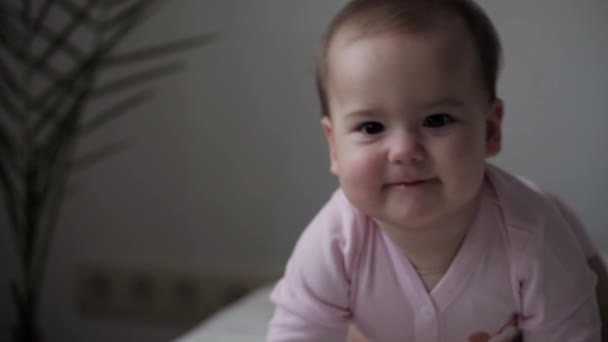 Bebé, concepto de la infancia - primer plano de sonriente feliz regordeta divertida cara gordito de ojos marrones del niño niña niña despierta. 9-12 meses bebé muecas, cierra los ojos se arrastra sentarse en la cama suave de color rosa en casa en interiores — Vídeos de Stock