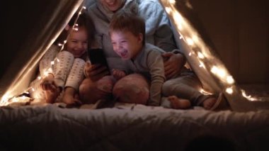 Otantik sevimli anne ve anaokulu çocukları evdeki çadırda kız ve erkek. Genç bir kadın gece vakti çadırda 2-4 yaşlarındaki çocuklarla akıllı telefon kullanıyor. Aile, Çocukluk, Annelik, Rahatlık ve Güvenlik kavramı