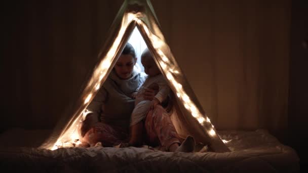 Authentic cute mom with chubby babe infant girl in tent at home. young woman use smartphone with kid 6-12 months old sit in wigwam at night. Family, Childhood, Motherhood, Comfort and Safety concept. — Stock Video