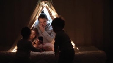 Otantik sevimli anne ve anaokulu çocukları evdeki çadırda kız ve erkek. Genç bir kadın gece vakti çadırda 2-4 yaşlarındaki çocuklarla akıllı telefon kullanıyor. Aile, Çocukluk, Annelik, Rahatlık ve Güvenlik kavramı