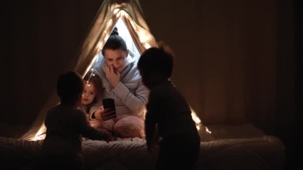 Authentic cute mom and preschool kids boy and girl in tent at home. young woman use smartphone with children 2-4 years old in wigwam at night. Family, Childhood, Motherhood, Comfort and Safety concept — Stock Video