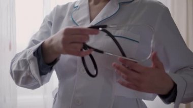 Tıp ve sağlık, pediatri, covid-19 konsepti - yakın plan genç kadın beyaz tıbbi önlük düğmesi tak ve pencerenin arka planına stetoskop tak. Hemşire ya da doktor kıyafeti. Ameliyat için hazırlık