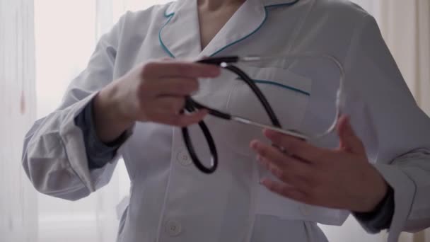 Medicína a zdraví, pediatrie, koncept covid-19 - zblízka mladá žena si na pozadí okna oblékla bílý knoflík lékařských šatů a stetoskop. sestře nebo doktorovi. příprava na operaci — Stock video