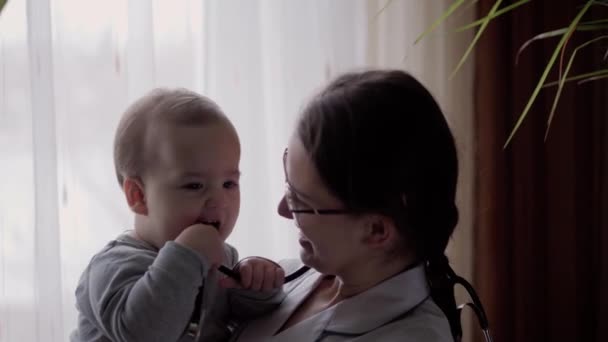 Medycyna i zdrowie, pediatria, koncepcja covid-19 - zbliżenie młoda kobieta pielęgniarka lub lekarz pediatra kaukaskiego wyglądu słowiańskiego trzymać niemowlę 8-12 miesięcy dziecko w ramionach na tle okna — Wideo stockowe