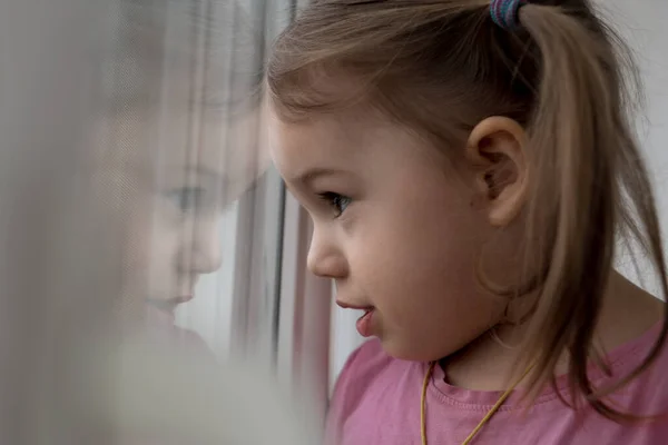 Kindertijd, eenzaamheid, isolatie, verdriet, hoop concepten - Een verdrietig schattig bezorgd Peuter baby meisje in roze met paardenstaarten kapsel kijkt door het raam. reflectie van het gezicht van het kleine lieve kind — Stockfoto