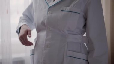 Tıp ve sağlık, pediatri, covid-19 konsepti - yakın plan genç kadın beyaz tıbbi önlük düğmesi tak ve pencerenin arka planına stetoskop tak. Hemşire ya da doktor kıyafeti. Ameliyat için hazırlık