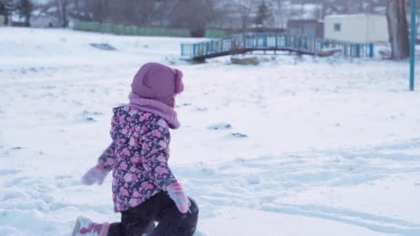 Zima, wakacje, gry, koncepcja rodziny - środkowy plan autentycznego małego przedszkola moll 3-4-letnia dziewczynka w fioletowym kolorze na pokrytej śniegiem łące bawi się śniegiem przed zachodem słońca. dziecko biegnie po śnieżnej okolicy — Wideo stockowe