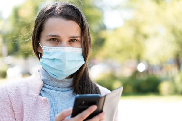 Bakım, bebekler, ilkbahar, cvid-19, karantina konsepti - Genç, uzun saçlı, Avrupalı, beyaz kadın tıbbi koruyucu maskeli, gün ortasında parkın arka planında akıllı telefon kullanıyor