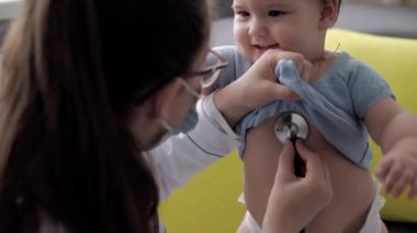 Tıp ve sağlık, pediatri, covid-19 konsepti - Kafkas Slav kökenli genç kadın hemşire ya da doktor çocuk doktorunu 8-12 aylık bebeği sarı gri kanepede muayene ediyor.
