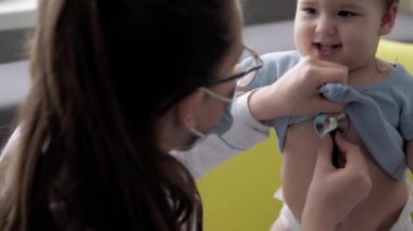 Tıp ve sağlık, pediatri, covid-19 konsepti - Kafkas Slav kökenli genç kadın hemşire ya da doktor çocuk doktorunu 8-12 aylık bebeği sarı gri kanepede muayene ediyor.