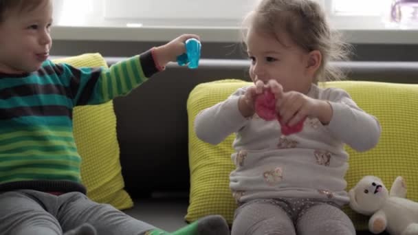 Αυθεντικά χεράκια μωρού με μπλε και κόκκινη γλίτσα. Κορίτσι και αγόρι προνήπιο μικρό τέντωμα Play-Doh στις πλευρές. Παιδί που παίζει πλαστελίνη. Μοντέρνο υγρό παιχνίδι κολλάει στα χέρια και τα δάχτυλα σε κίτρινο γκρι καναπέ — Αρχείο Βίντεο