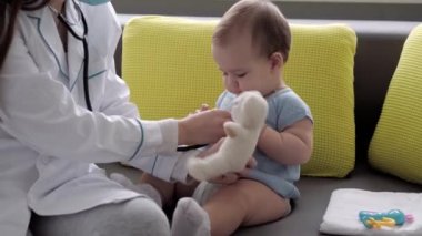 Tıp ve sağlık, pediatri, covid-19 konsepti - Kafkas Slav kökenli genç kadın hemşire ya da doktor çocuk doktorunu 8-12 aylık bebeği sarı gri kanepede muayene ediyor.