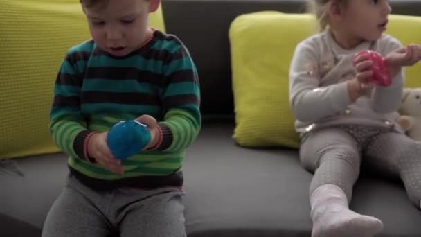 Autentické dětské ruce s modrým a červeným slizem. Dívka a chlapec předškolního věku natahují Play-Doh na stranu. dítě hrající si na plastikové těsto. Módní tekutá hračka se lepí na ruce a prsty na žluté šedé pohovce — Stock video