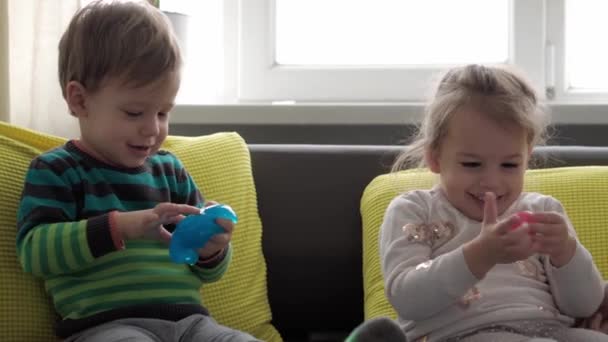 Äkta babyhänder med blått och rött slem. Flicka och pojke förskola mindre sträcker Play-Doh till sidor. Grabben spelar plastdeg. Trendig flytande leksak fastnar på händer och fingrar på gul grå soffa — Stockvideo