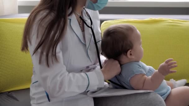 Medicina y salud, pediatría, covid-19 concepto primer plano joven enfermera o médico pediatra de etnia eslava caucásica examina bebé de 8-12 meses en el sofá gris amarillo ventana opuesta — Vídeo de stock