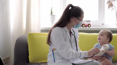 Tıp ve sağlık, pediatri, covid-19 konsepti - Kafkas Slav kökenli genç kadın hemşire ya da doktor çocuk doktorunu 8-12 aylık bebeği sarı gri kanepede muayene ediyor.
