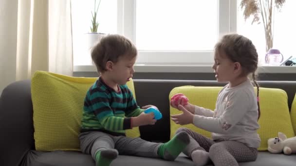 Äkta babyhänder med blått och rött slem. Flicka och pojke förskola mindre sträcker Play-Doh till sidor. Grabben spelar plastdeg. Trendig flytande leksak fastnar på händer och fingrar på gul grå soffa — Stockvideo