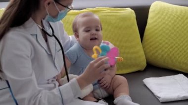 Tıp ve sağlık, pediatri, covid-19 konsepti - Kafkas Slav kökenli genç kadın hemşire ya da doktor çocuk doktorunu 8-12 aylık bebeği sarı gri kanepede muayene ediyor.