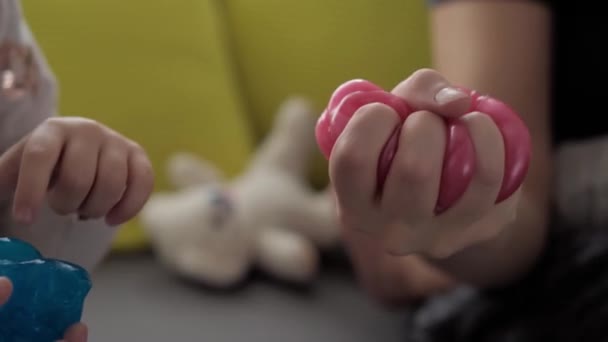 Ruce s modrým a červeným slizem zblízka. Dívka a chlapec předškolního věku natahují Play-Doh na stranu. děti hrající si na plastikové těsto. Módní tekutá hračka se lepí na ruce a prsty na žluté šedé pohovce — Stock video