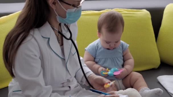 医学与健康、儿科、 covid-19概念- -近视的年轻女护士或医生、高加索斯拉夫裔儿科医生在对面的黄灰色沙发上检查8-12个月大的婴儿 — 图库视频影像