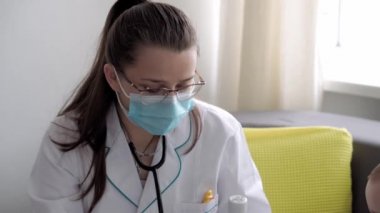 Tıp ve sağlık, pediatri, covid-19 konsepti - Kafkas Slav kökenli genç kadın hemşire ya da doktor çocuk doktorunu 8-12 aylık bebeği sarı gri kanepede muayene ediyor.