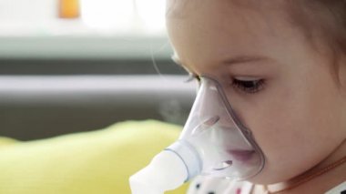 3-4 yaşlarında, sarı gri kanepenin üzerinde astım spreyi maskesi takmış, anaokulu öğrencisi bir çocuk. Bebek nebulizörle solunum terapisi görüyor. ilaç ve sağlık, pediatri, covid-19 konsepti