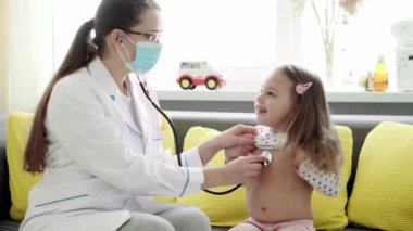 Kafkas Slav kökenli genç kadın hemşire veya doktor çocuk doktorunun orta dereceli planı, sarı gri kanepede 3-4 yaşındaki anaokulu öğrencisi kızı muayene ediyor. ilaç ve sağlık, pediatri, covid-19 konsepti